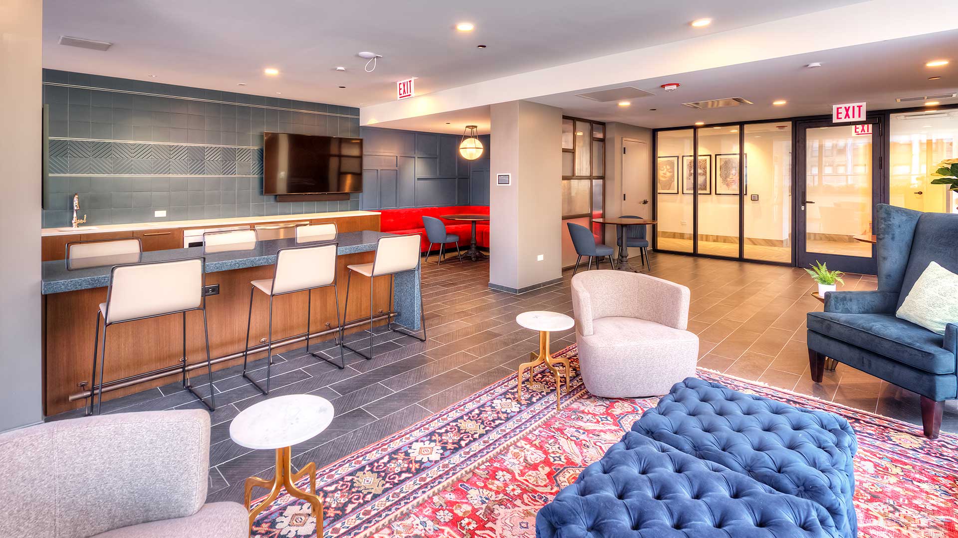 There is an ottoman and a few club chairs on a nice rug in the foreground. A bar and television are off to the left with bench seating extending back into a far corner. To the far right is a glass wall and the entrance to the clubroom.