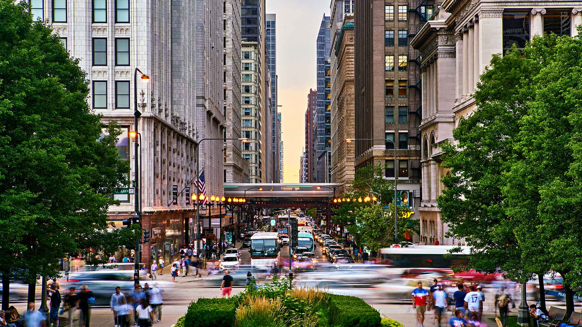 Looking down a city street in Chicago. The elevated train passes over the street a block ahead. There are people walking all over and cars driving.