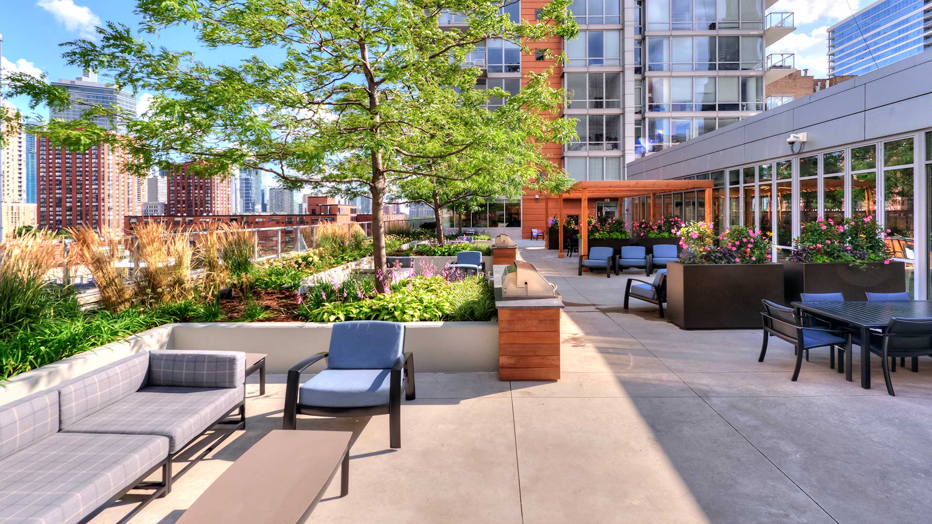 Looking down the outdoor terrace at Burnham Pointe. Along the left are various outdoor furniture pieces and potted plants, the city seen in the distance. Along the right is more seating and windows that look into the entertainment lounge.
