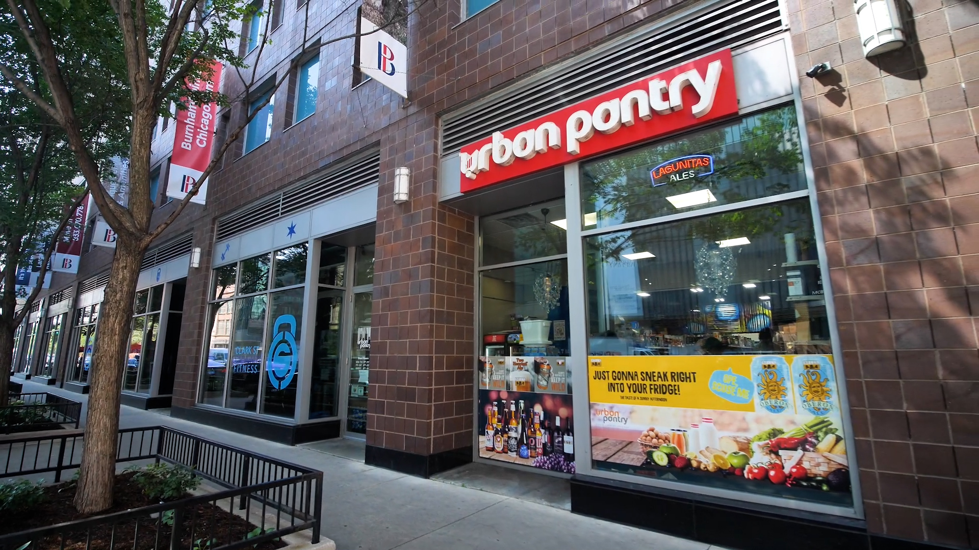 Standing on the sidewalk looking angled at the storefront to Urban Pantry at Burnham Pointe. There are ads in the windows and the shop is seen inside.